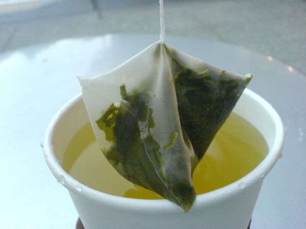 Pyramidal tea bag suspended from its string, filled with wet green tea leaves, over a white paper cup filled with green tea