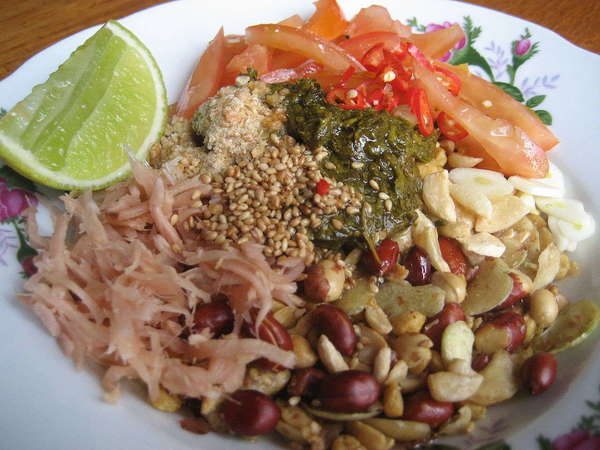 A plate with a lime slice, pickled tea leaves, various beans and seeds, and other veggies