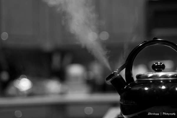 Black and white photo of dark teakettle on stove emitting steam, in kitchen setting
