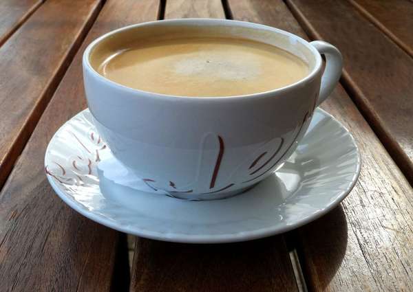Cup of coffee with milk, foamy appearance, in saucer, on dark wooden table with space between slats