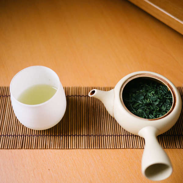 Cup on left with pale green tea, traditional Japanese teapot on right with spent (steeped) green tea leaves, on a bamboo mat against a wood-colored background