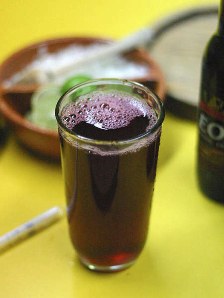 Glass filled with dark purple-red drink, a bowl in the background and a bottle on the right, background blurred, on a yellow surface