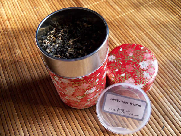 Red floral print tin on bamboo mat, containing curly loose-leaf black tea