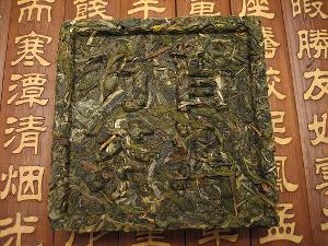 A pu-erh cake pressed into a square, with Chinese characters printed on it, with a dull green leaf color