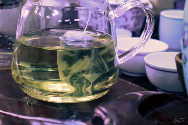 Large tea bag inside a glass teapot, the liquid showing a pale yellow color, with empty white teacups in the background