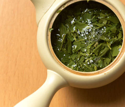 Overhead view of loose-leaf green tea, flaky and broken in appearance, steeping in a traditional Japanese teapot with lid open