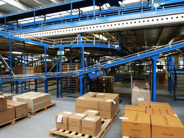 Interior of warehouse showing blue-painted conveyor belts and numerous boxes set out on wooden pallets