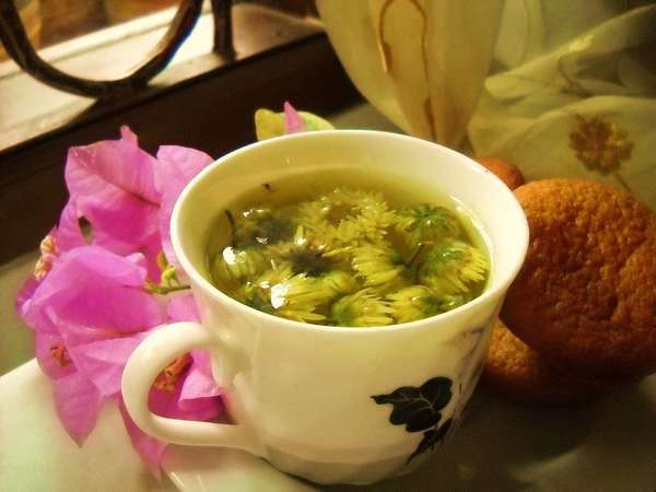 Cup full of Chrysanthemum blosssoms steeping in water, next to cookies and fake flowers