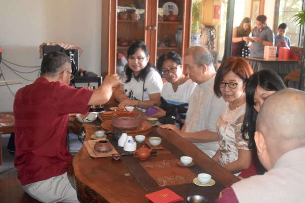 Six people gathered around a table on which a man is brewing and pouring tea for them