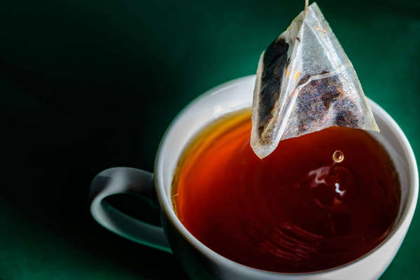 Paper pyramid tea bag being lifted from a white cup filled with rich red-colored tea, on a green background