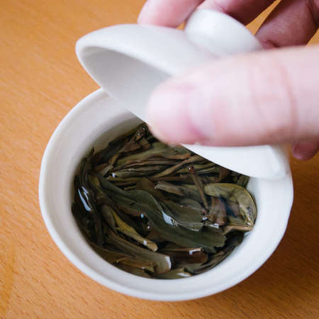 White gaiwan with dark-olive whole tea leaves, hand holding the lid above