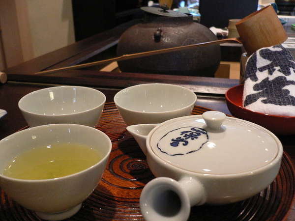 Brewed green tea in a broad teacup to the left, a traditional Japanese teapot to the right, in a tea house setting
