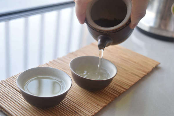 Hand pouring pale tea into tiny teacups on a bamboo mat, in plain, bright white surroundings