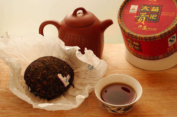 Compressed tea cake in paper wrapping, dark brown brewed cup of tea, clay teapot, and red cylindrical tea packaging