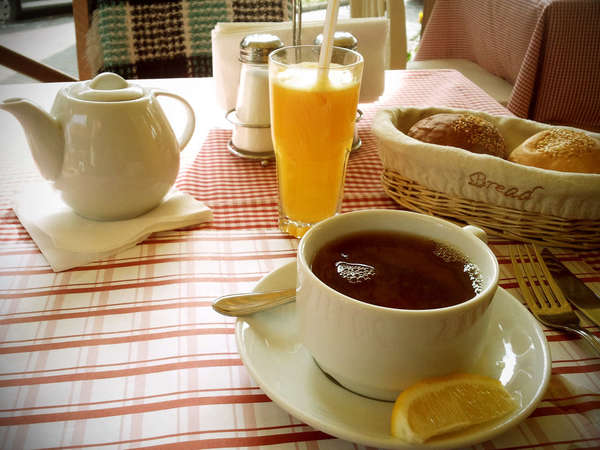 Cup of black tea in simple white cup and saucer with slice of lemon, basket of rolls, cup of orange juice, and teapot, on red and white tablecloth