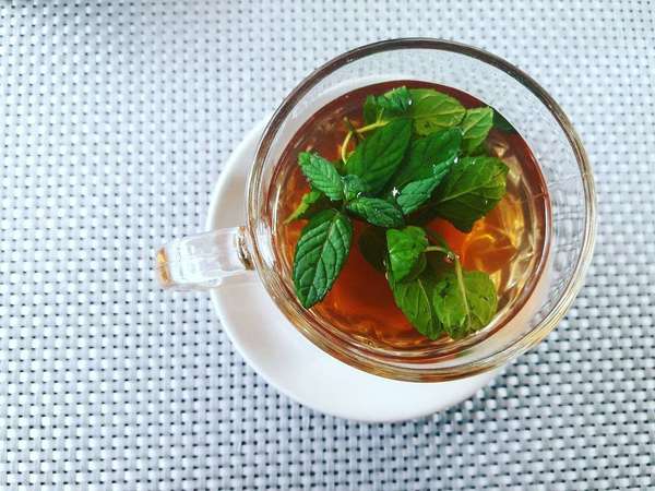 Overhead view of glass teacup containing tea and fresh spearmint leaves, on a white saucer, on a grayish mesh background