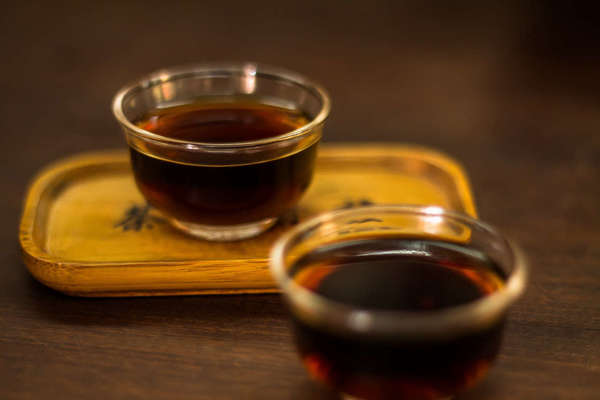 Two small glass cups of very dark tea, one on a tray, on a dark wooden surface