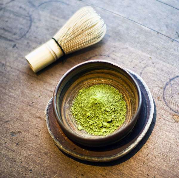 Black and brown ceramic bowl filled with pale green tea powder, bamboo whisk on side, on wooden surface