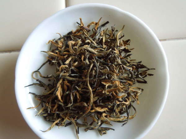 Loose-leaf tea with black and orange, long, wiry leaves, in white dish on tile background