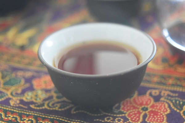 Ceramic cup, white inside, black outside with dark tea liquor, on ornate tablecloth with red flowers, green leaves, gold trim againsst purple background