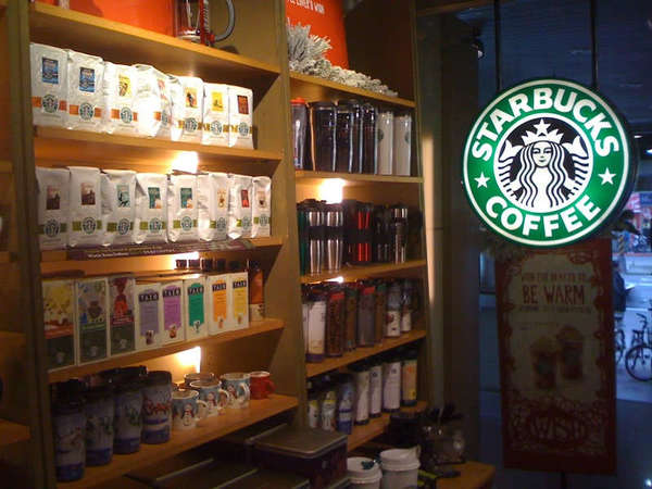 Side wall of Starbucks store showing lit-up Starbucks sign, and boxes of tea and drink travel mugs for sale