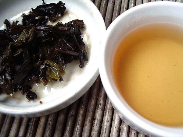 Cup of pale-golden tea on right, dark green steeped leaves, crinkly in texture, in a dish to the left