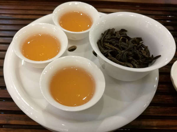 Plate with wet dark tea leaves in a bowl, and three cups of golden-colored tea, on wooden tray