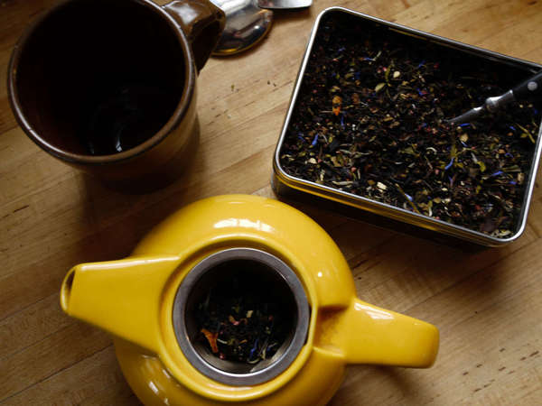 Colorful, loose-leaf flavored tea blend in a tin top right, the same tea steeping in a yellow teapot below, viewed from above, mug top left