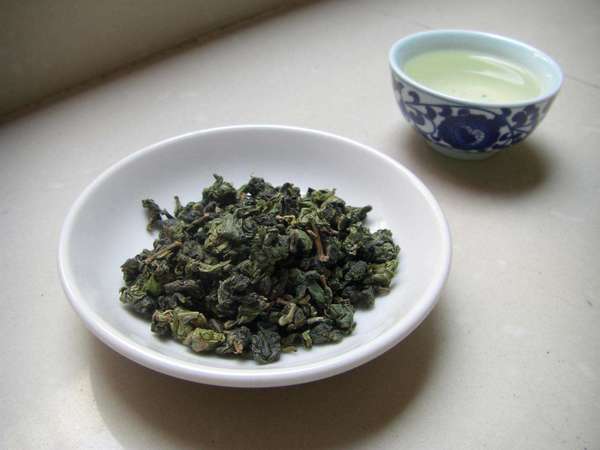 Vibrant green, tightly-rolled tea leaves in a white dish, a cup filled with pale green brewed tea behind it
