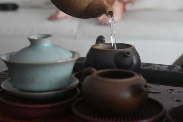 Tea being poured into a small ceramic teapot, a gaiwan on the left, and another open teapot in the foreground