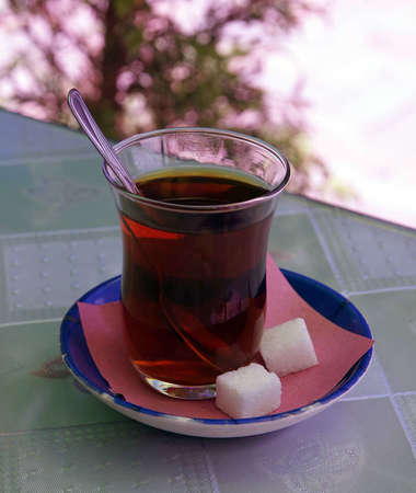 Reddish-colored black tea in glass teacup with spoon, on blue saucer with two sugar cubes