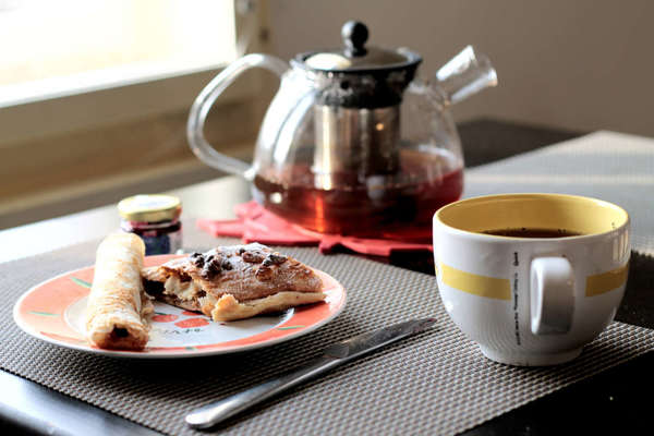 Black tea in teapot with built-in infuser, cup of tea, and crepes on a plate