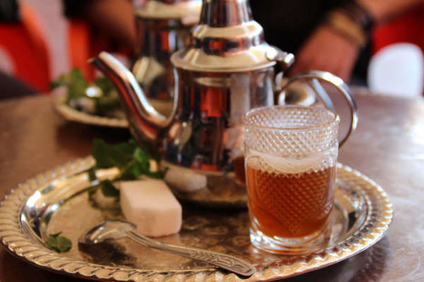 Shiny metal teapot with a glass of amber-colored tea in the foreground, on a shiny platter with a spoon and large sugar cube