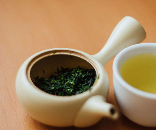 Japanese tea pot filled with steeped green tea leaves, cup full of green tea on the right